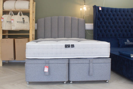 Gray mattress set with tufted headboard and blue sofa in a room with storage shelves.